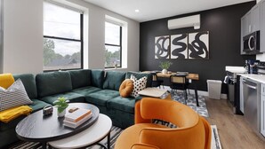 Una sala moderna con un sofá seccional verde, una mesa de centro redonda y un sillón naranja. Se observa un área de cocina con un microondas y un lavabo.