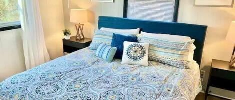 A bedroom with a patterned bedspread, a blue headboard, and a window with blinds.
