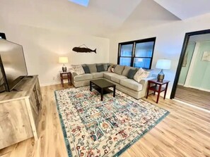 Una sala de estar con un sofá modular, una mesa de centro, un tapete estampado, una decoración de peces en la pared y un televisor sobre un mueble de madera.