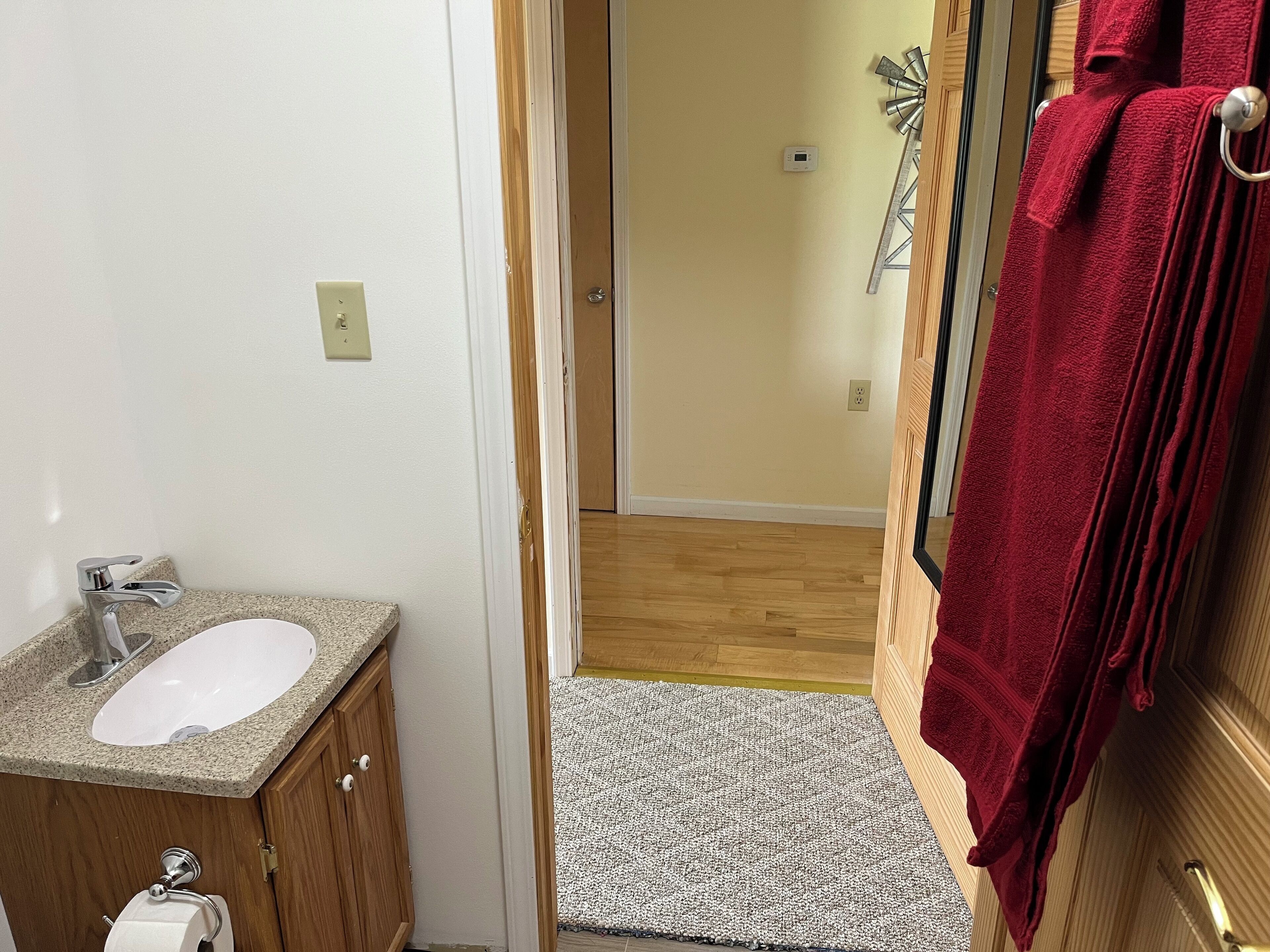 Un baño con lavamanos blanco, mueble de madera y una toalla roja colgada en la puerta.