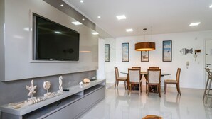Een moderne woonkamer met een flatscreen televisie, een eethoek met een tafel en stoelen, en een grote spiegel aan de muur.