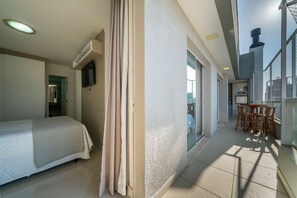 A hotel room with a bed, a television, a balcony with a table and chairs, and a view of a building. - Residencial Florenza (Bombinhas)