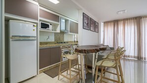 Een kleine keuken met een koelkast, magnetron en kookplaat, een eethoek met barstoelen, en een groot raam met gordijnen.