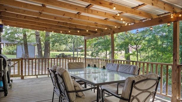 A covered outdoor patio with a glass-top table and cushioned chairs, surrounded by wooden railings and trees.