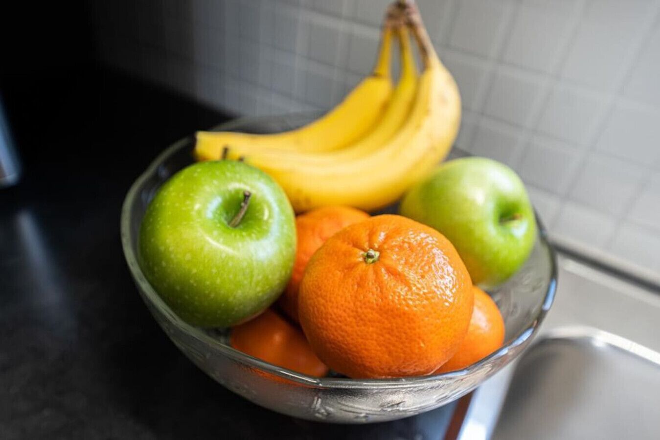 A glass bowl containing green apples, an orange, and a bunch of bananas on a kitchen counter.