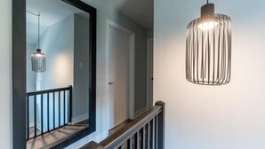 A hallway with a large mirror, a wooden railing, and a hanging pendant light. - Outdoor Sauna|Hot Tub|Playroom|Port Dallhousie (St. Catharines)