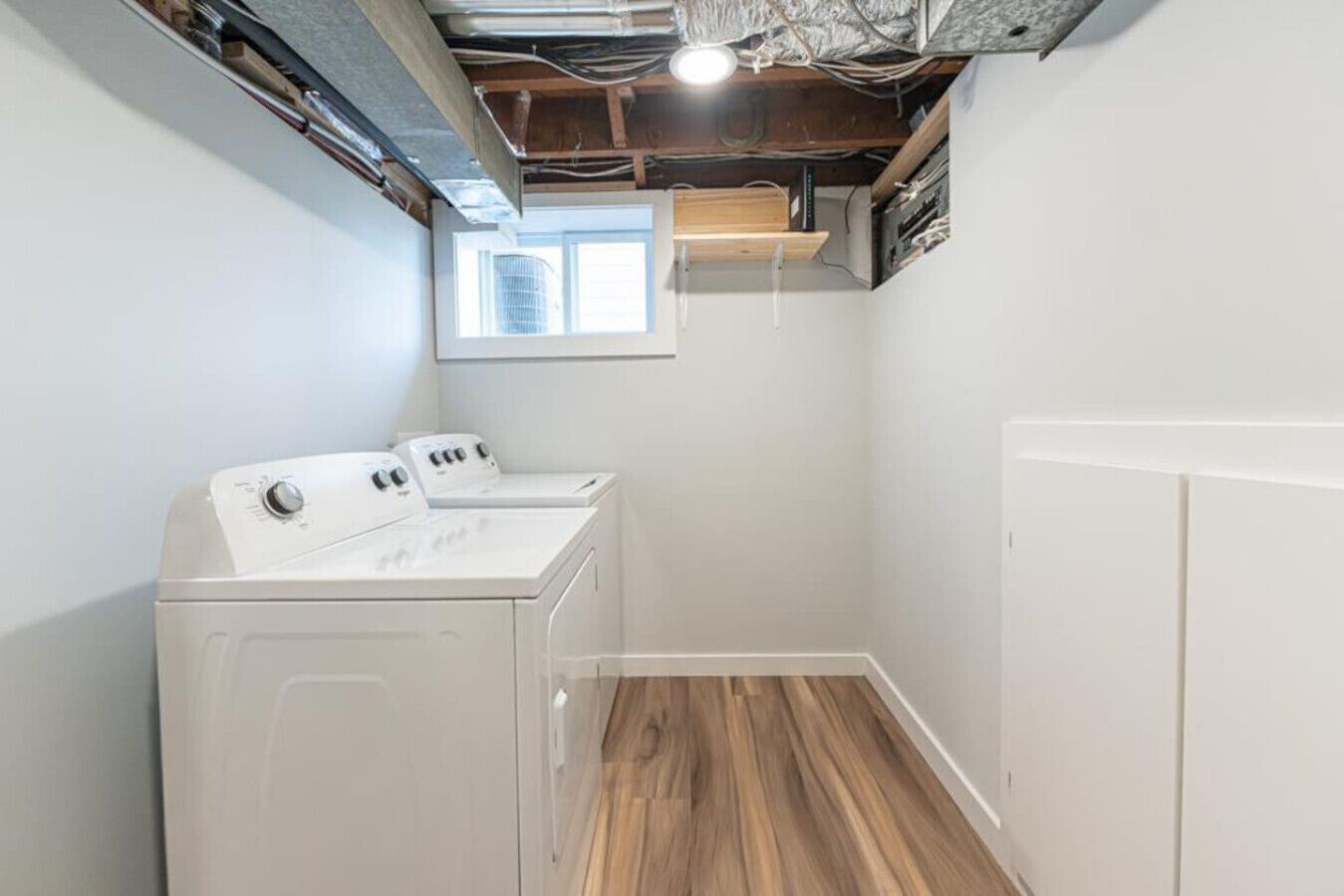 A laundry room with two stacked washers and dryers, wooden flooring, and a window providing natural light.