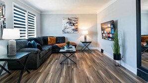 A modern living room with a dark-colored sofa, a coffee table, and a TV mounted on the wall. - Outdoor Sauna|Hot Tub|Playroom|Port Dallhousie (St. Catharines)