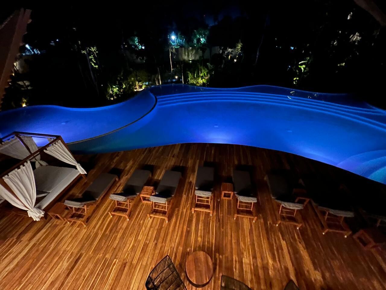 A wooden deck with lounge chairs and a small table, surrounded by a pool with blue lighting.