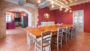Un comedor espacioso con una larga mesa de madera y sillas blancas, una cocina al fondo, y vigas de madera a la vista en el techo.