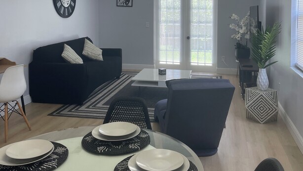 Una sala de estar moderna con comedor, que incluye una araña, un sofá negro, una mesa de comedor blanca con individuales negros y un ventanal con persianas.