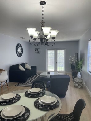 Un salon moderne avec un coin repas, comprenant un lustre, un canapé noir, une table à manger blanche avec des sets de table noirs et une grande fenêtre avec des stores.