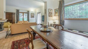 A spacious living room with a sofa, dining table, and chairs. There is a chandelier, a window with curtains, and a radiator. - Santa Colomba House, Siena, Nature And Pool (Monteriggioni)