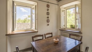 Un coin repas avec une table et des chaises en bois, deux fenêtres donnant sur un toit recouvert de tuiles et sur de la verdure, et un radiateur placé sous les fenêtres.