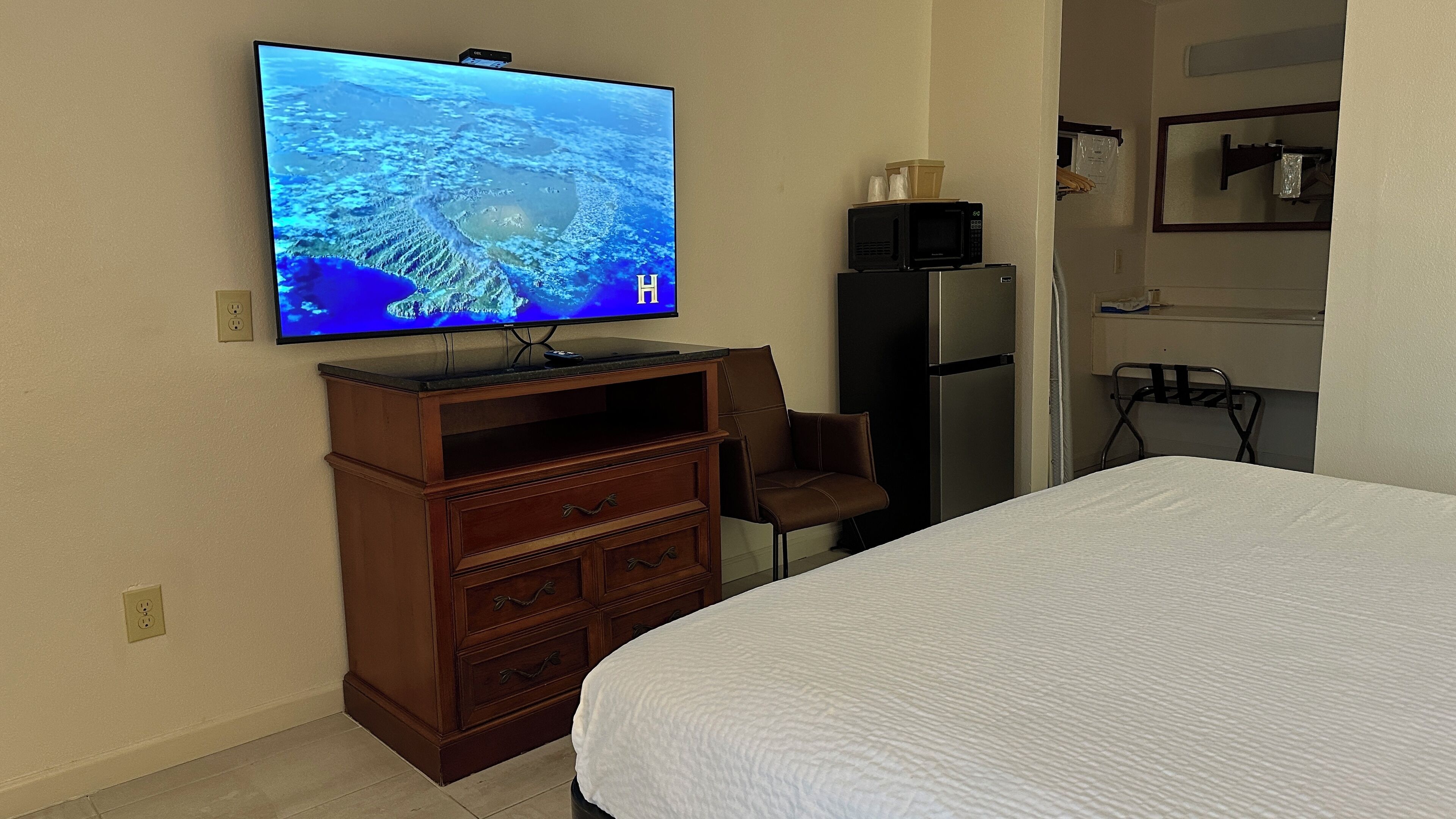 A hotel room with a bed, a TV mounted on the wall, a wooden dresser, a refrigerator, and a microwave.