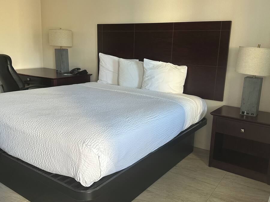 Una habitación de hotel con una cama grande, un cabecero oscuro, dos mesitas de noche con lámparas y una silla.