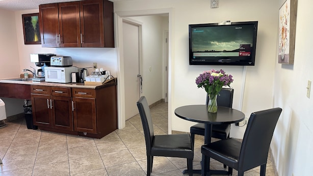 A compact kitchen with a microwave, coffee maker, and cabinets. There's a dining area with a table, chairs, and a vase of flowers. A TV is mounted on the wall displaying a weather forecast. - Carom Inn Denham Springs-Baton Rouge (Denham Springs)