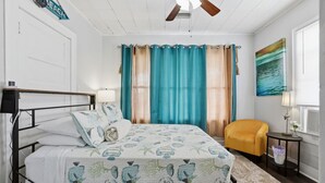 Un dormitorio con cama, una silla, un ventilador de techo, cortinas y un cuadro en la pared.