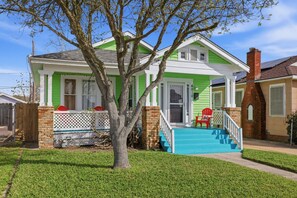 Uma casa de dois andares com fachada verde e branca, um alpendre com uma cadeira vermelha e uma escadaria azul que leva à entrada.