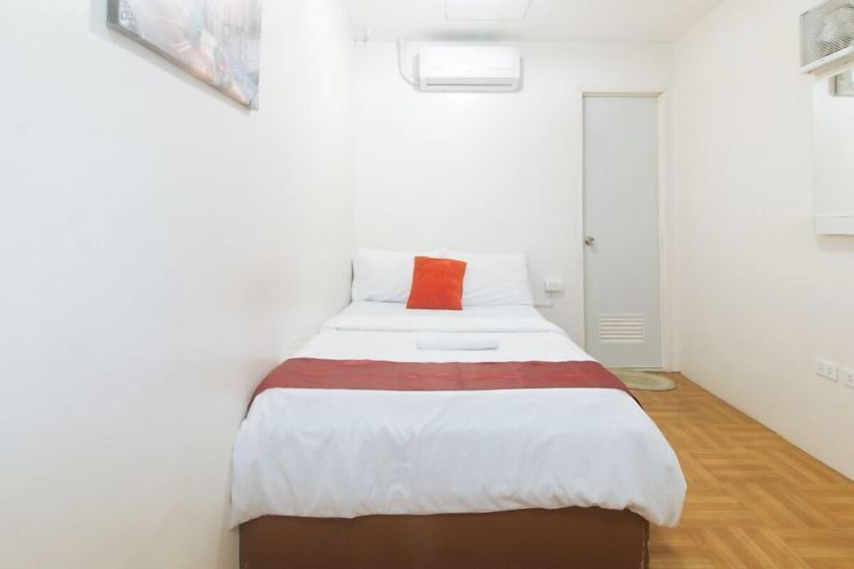 Un lit simple avec une couvre-lit blanche et rouge, un oreiller blanc et un oreiller décoratif rouge. Un climatiseur blanc est fixé au mur au-dessus du lit, et une porte se trouve sur le côté droit.