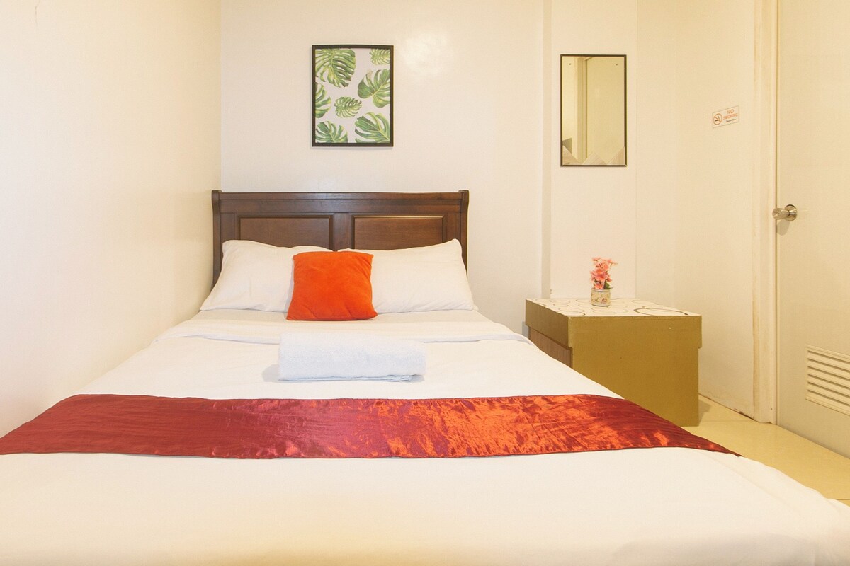 A neatly made bed with white and red bedding, a wooden headboard, a small nightstand with a vase of flowers, and a framed plant on the wall.