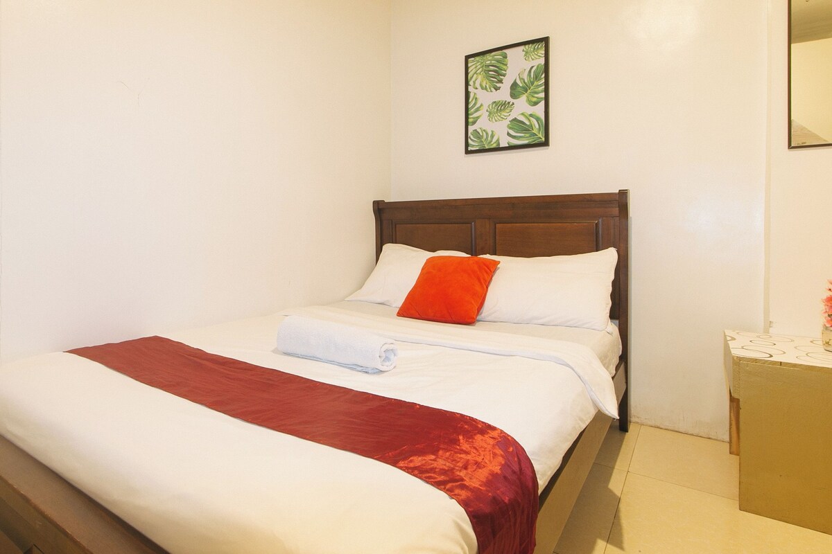 A neatly made bed with white and red linens, a wooden headboard, and a single orange pillow.