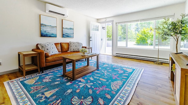 Ein Wohnzimmer mit einem braunen Ledersofa, einem Holztisch und einem bunten Teppich. Zwei Wandairconditioner und ein großes Fenster mit Blick auf Grünpflanzen sind vorhanden.