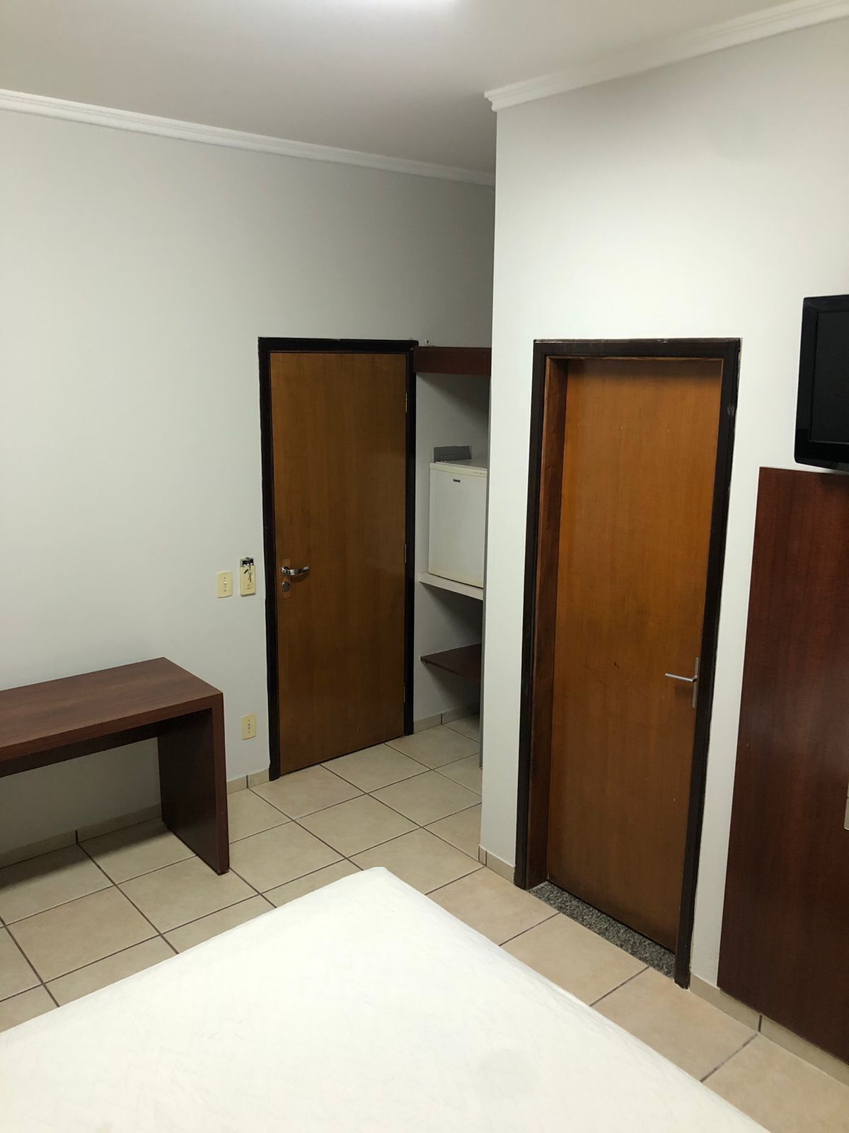 Quarto com cama, um banco de madeira, duas portas e uma televisão sobre um suporte.