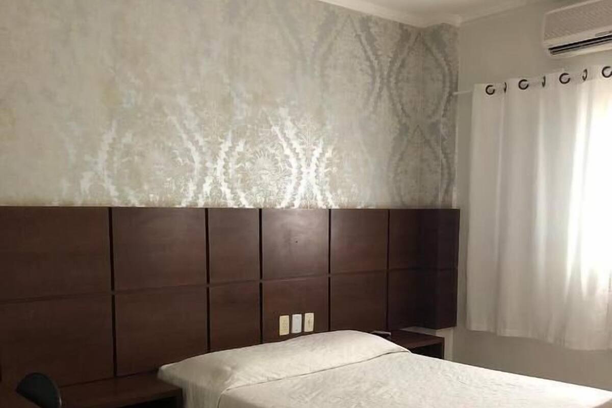 A hotel room with a large bed, a desk, and a chair. There is a wall-mounted air conditioning unit and a curtain on the window.