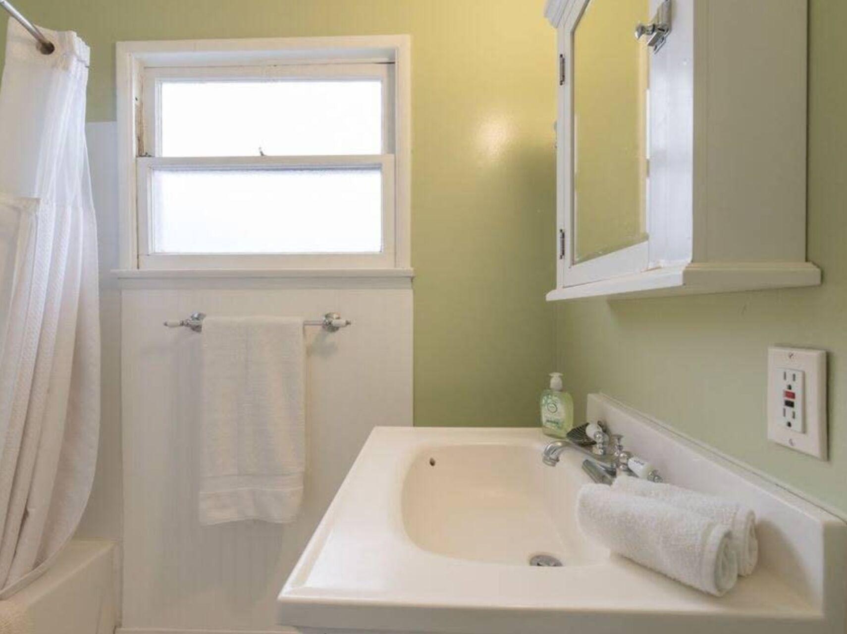 A bathroom with a white sink, a window above the mirror, and a towel hanging on the door.