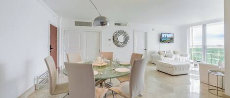 Un salon moderne avec une table à manger en verre, des canapés blancs et une grande fenêtre donnant sur la ville.