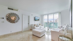 A spacious living room with a large window, a white sofa set, a round mirror, and a television on the wall. - Amazing Beachside Condo (Hollywood)