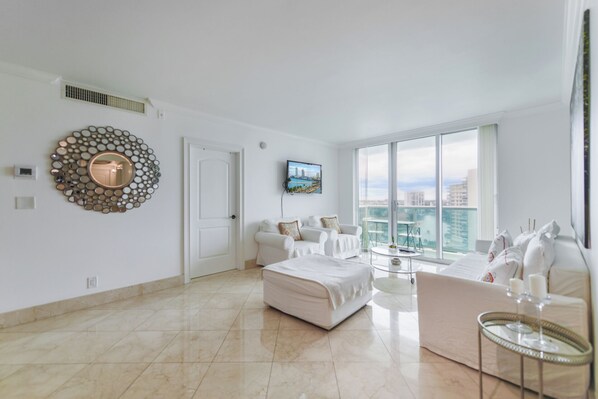 A spacious living room with a large window, a white sofa set, a round mirror, and a television on the wall. - Tides on Hollywood - Walkable to the Beach - We Host (Hollywood)