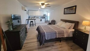 Quarto com cama, cômoda, televisão e cozinha compacta ao fundo.