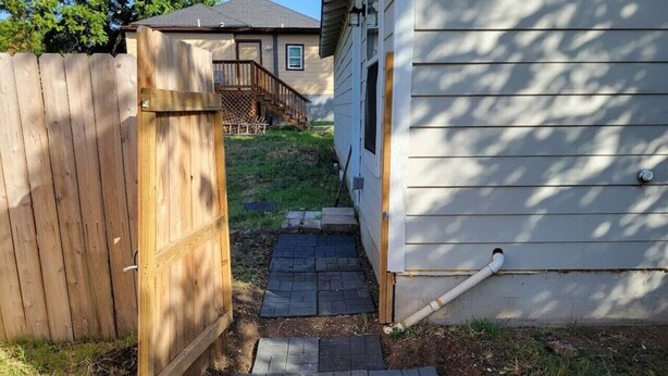 Uma trilha leva a uma casa com cerca de madeira de um lado e parede externa branca do outro.