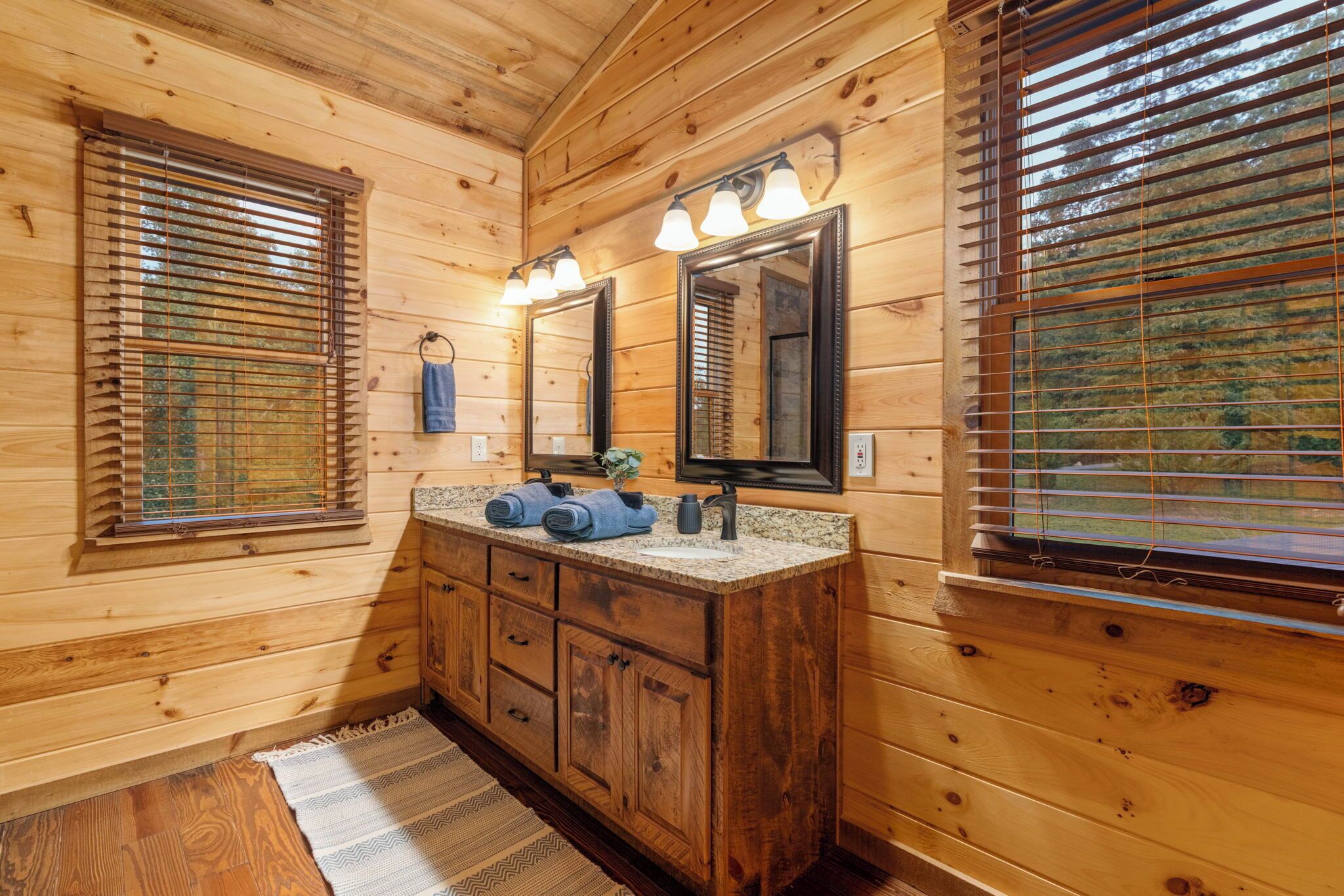 A wooden cabin bathroom with a granite countertop, wooden cabinets, a sink, and two mirrors.