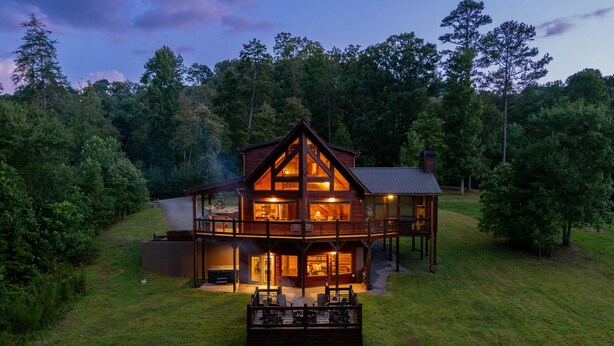 A two-story wooden cabin with a balcony, surrounded by a grassy yard and trees. - River Front! Pool Table! Riverfront Fire Pit! Hot Tub! BlueRidgeScenicRailway! (Blue Ridge)