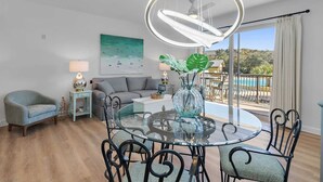 Ett modernt vardagsrum med ett matbord i glas, svarta metallstolar, en grå soffa, ett litet sidobord med en lampa och ett stort fönster med utsikt över en pool och grönområden.