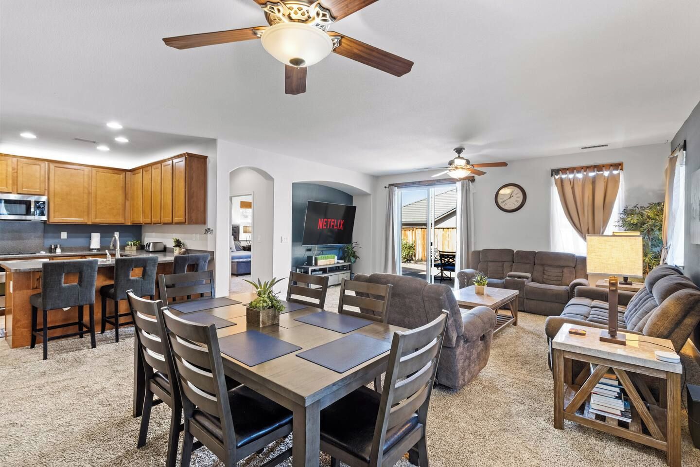 A modern kitchen with wooden cabinets, a dining area with a table and chairs, a living room with a sofa and a TV displaying 'NETFLIX,' and a ceiling fan.