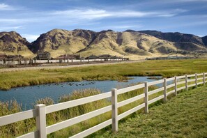 A river with a white fence in the foreground, a grassy field, and mountains in the background.