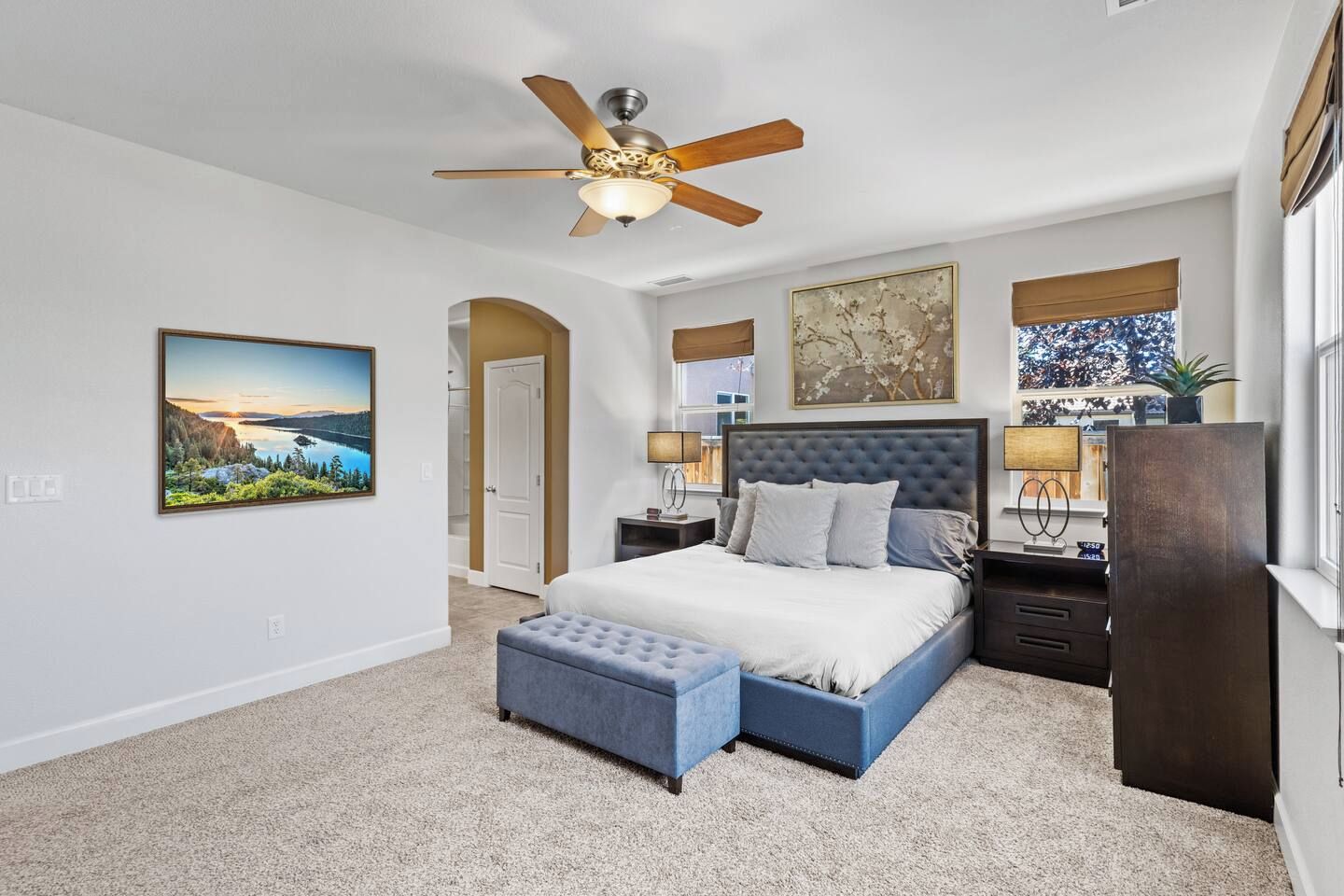 A bedroom with a bed, a nightstand, a lamp, a ceiling fan, a painting, and a window with a view.
