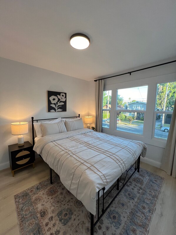 Ein Schlafzimmer mit Bett, Nachttischen, einer Lampe, einem Fenster mit Vorhängen und Blick nach draußen.