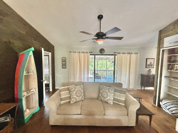 A living room with a beige sofa, patterned pillows, a ceiling fan, and a view through a glass door to an outdoor patio. - Our Anchor - close to the beach! (St. George Island)