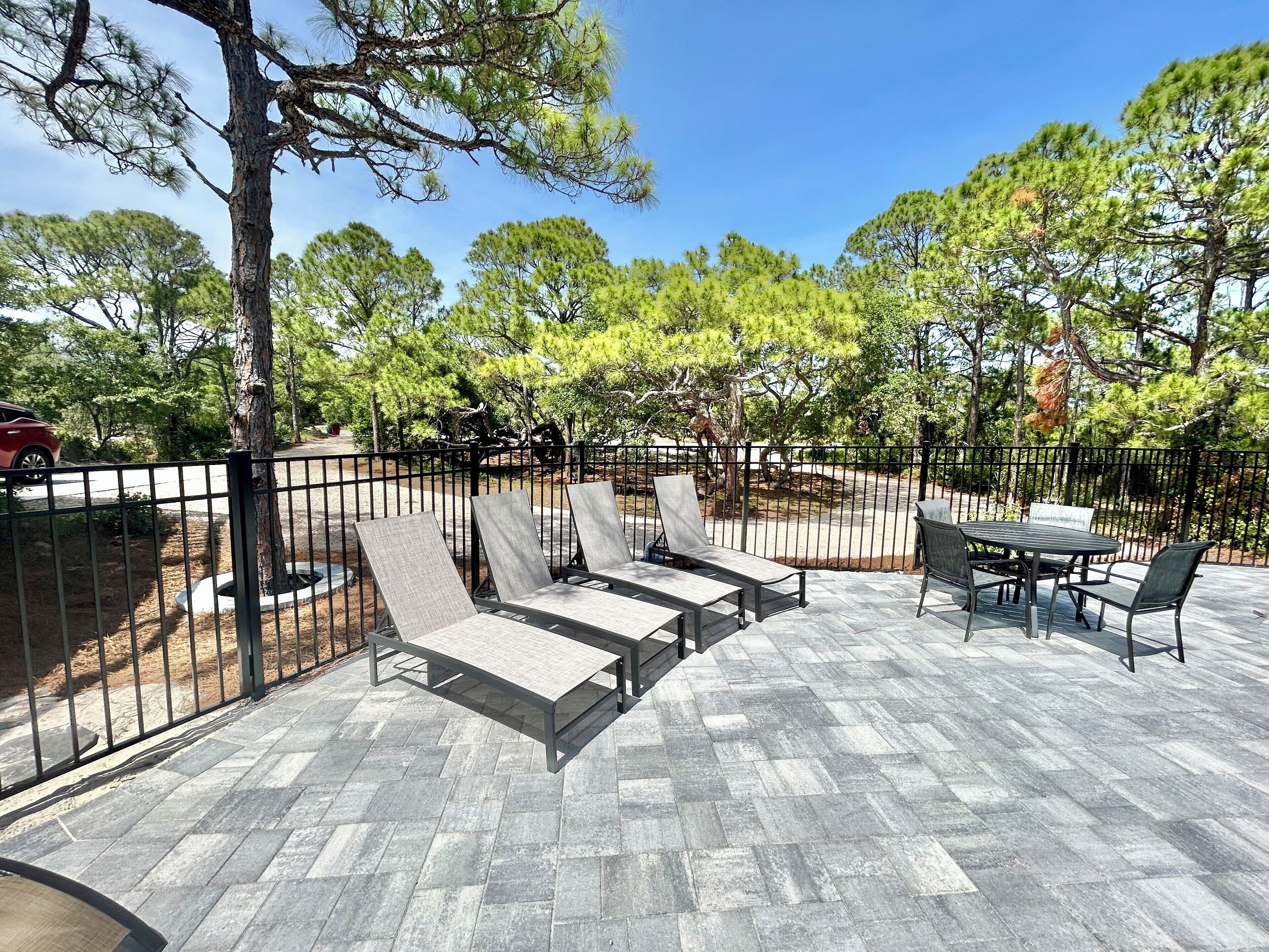 An outdoor patio with lounge chairs and a table, surrounded by a metal fence and trees.