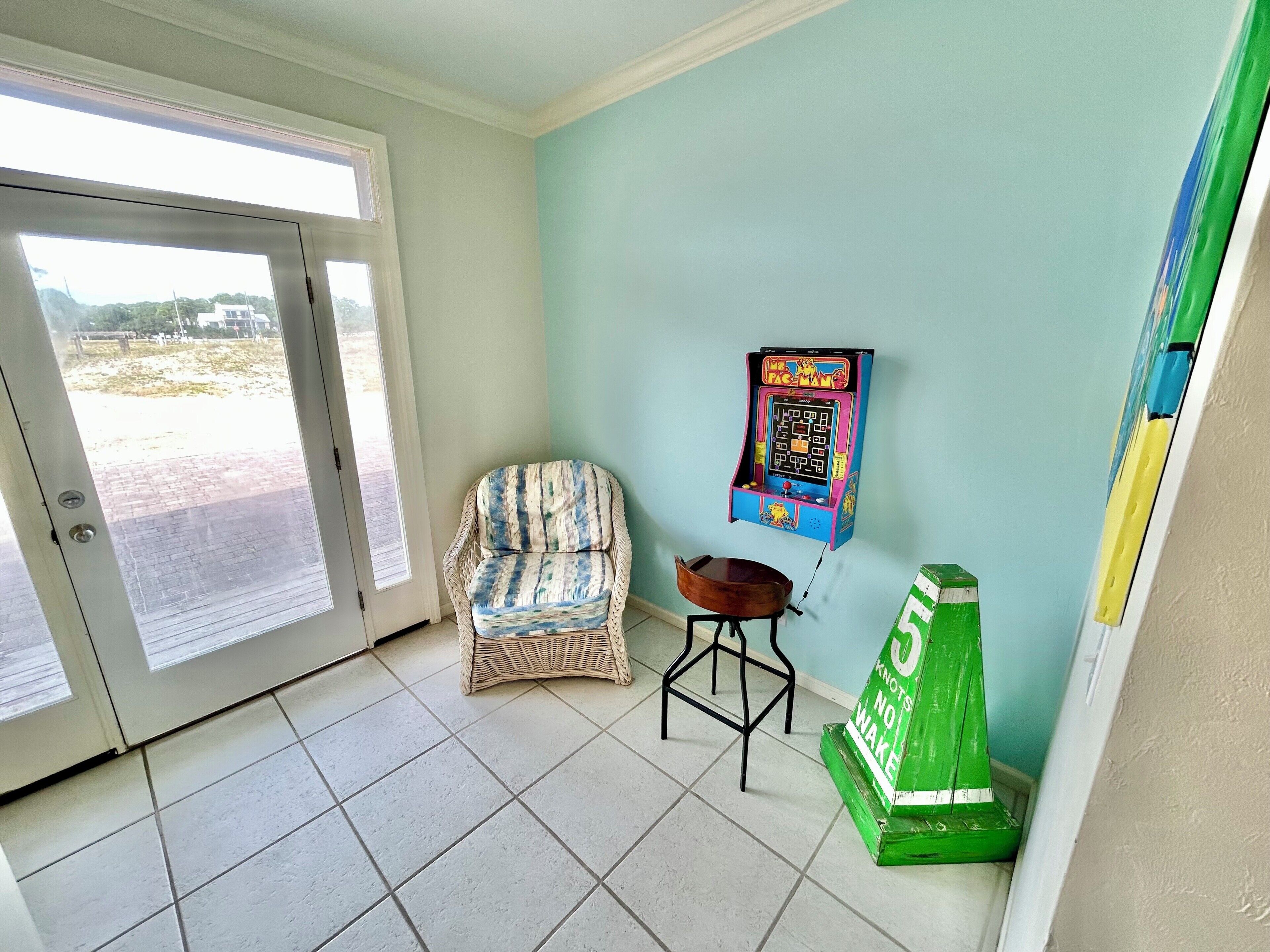 Una habitación con una puerta que da al exterior, una silla de mimbre, un taburete pequeño y una máquina arcade de Pac-Man.