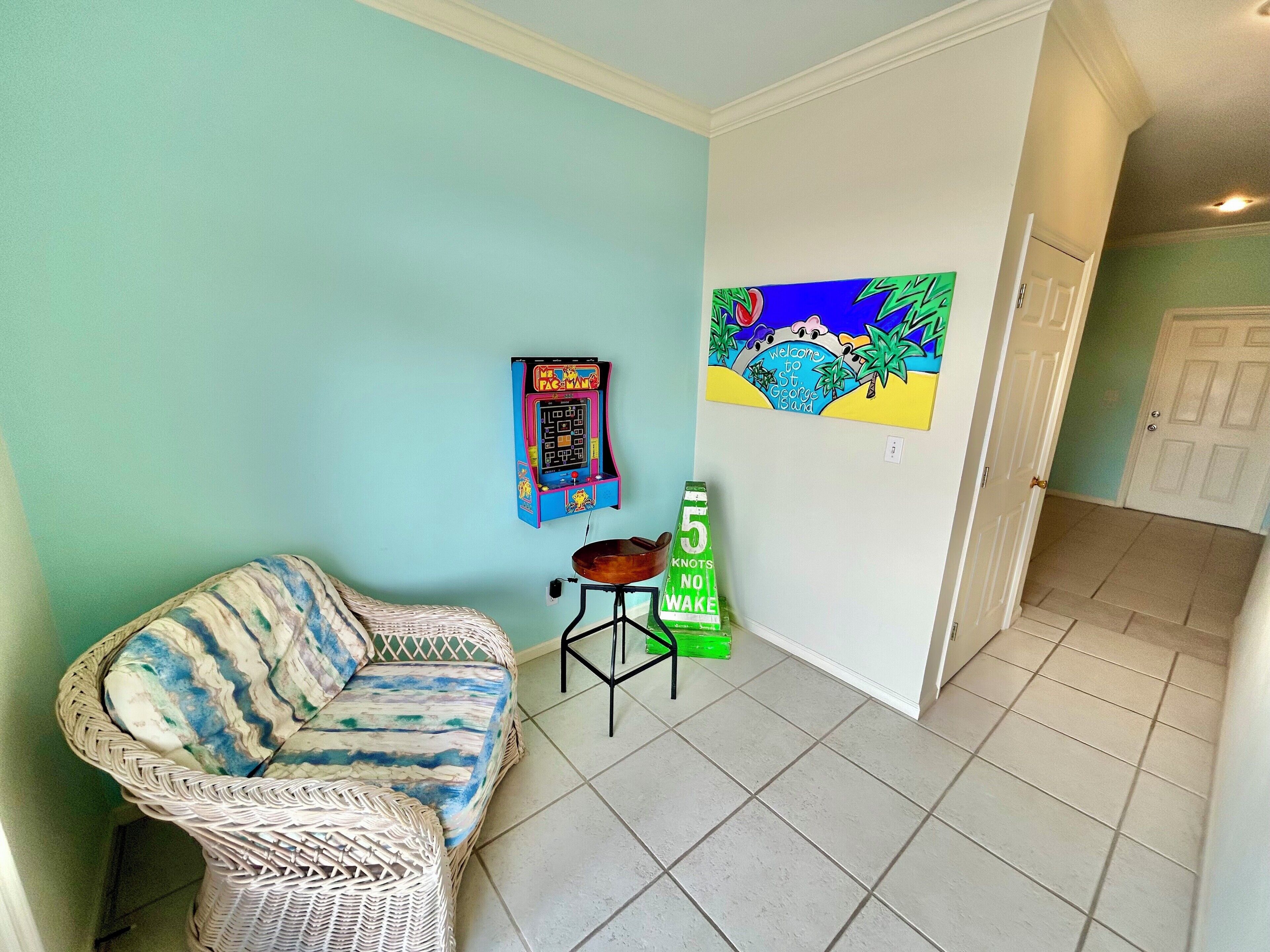 A room with a wicker sofa, a small table, a Pac-Man arcade game, a painting, and a green sign.