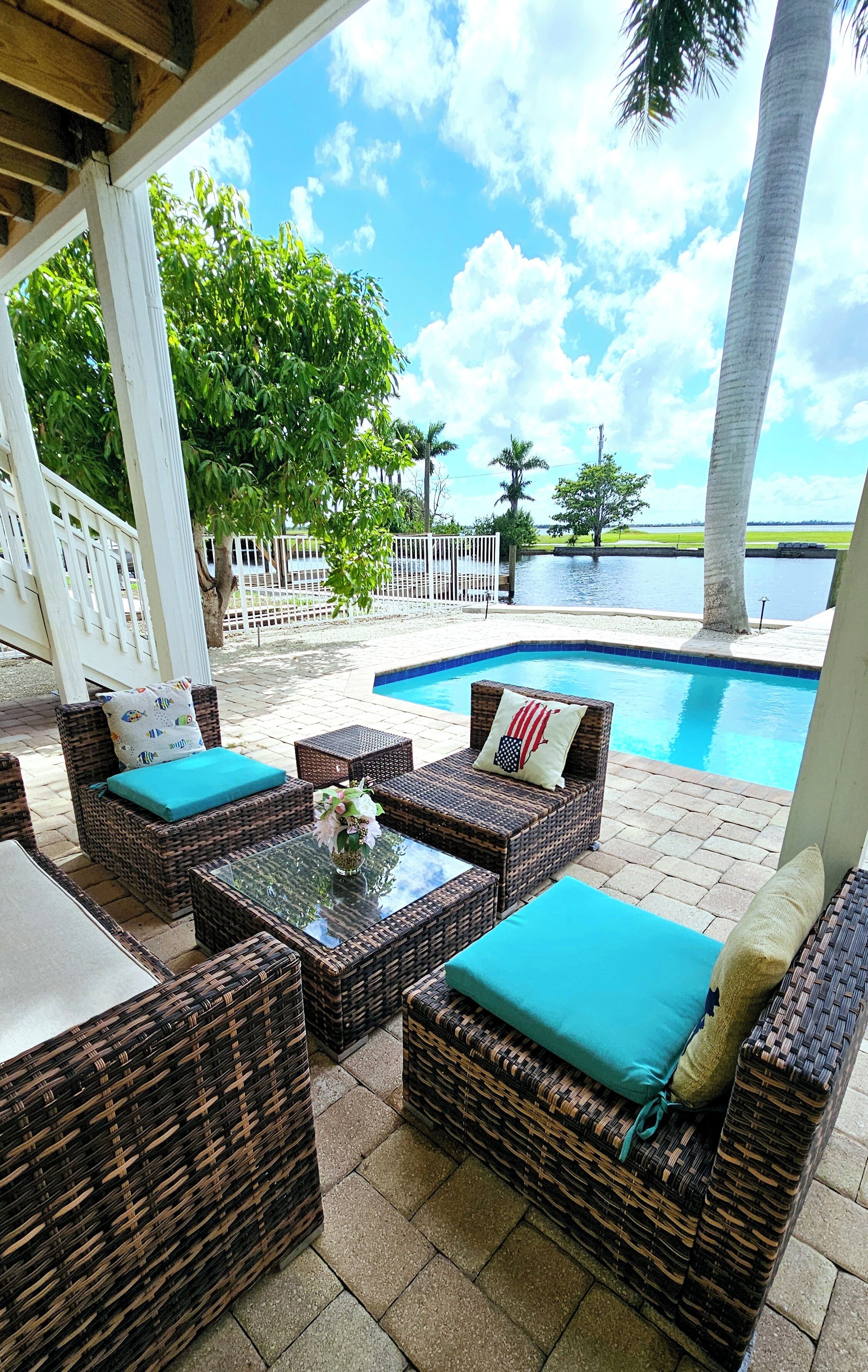 A covered outdoor seating area with wicker furniture and a glass table, overlooking a swimming pool and palm trees.