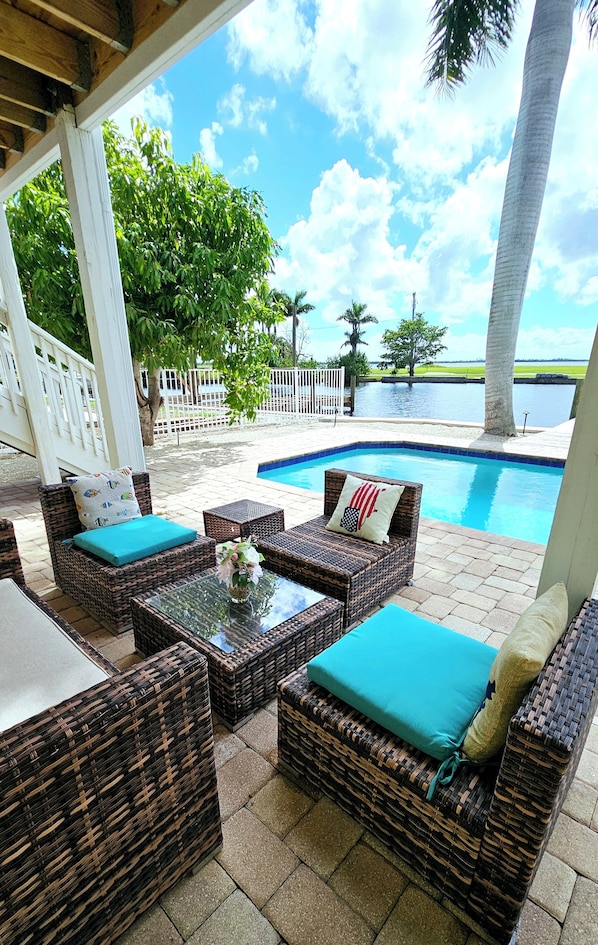 A covered outdoor seating area with wicker furniture and a glass table, overlooking a swimming pool and palm trees.