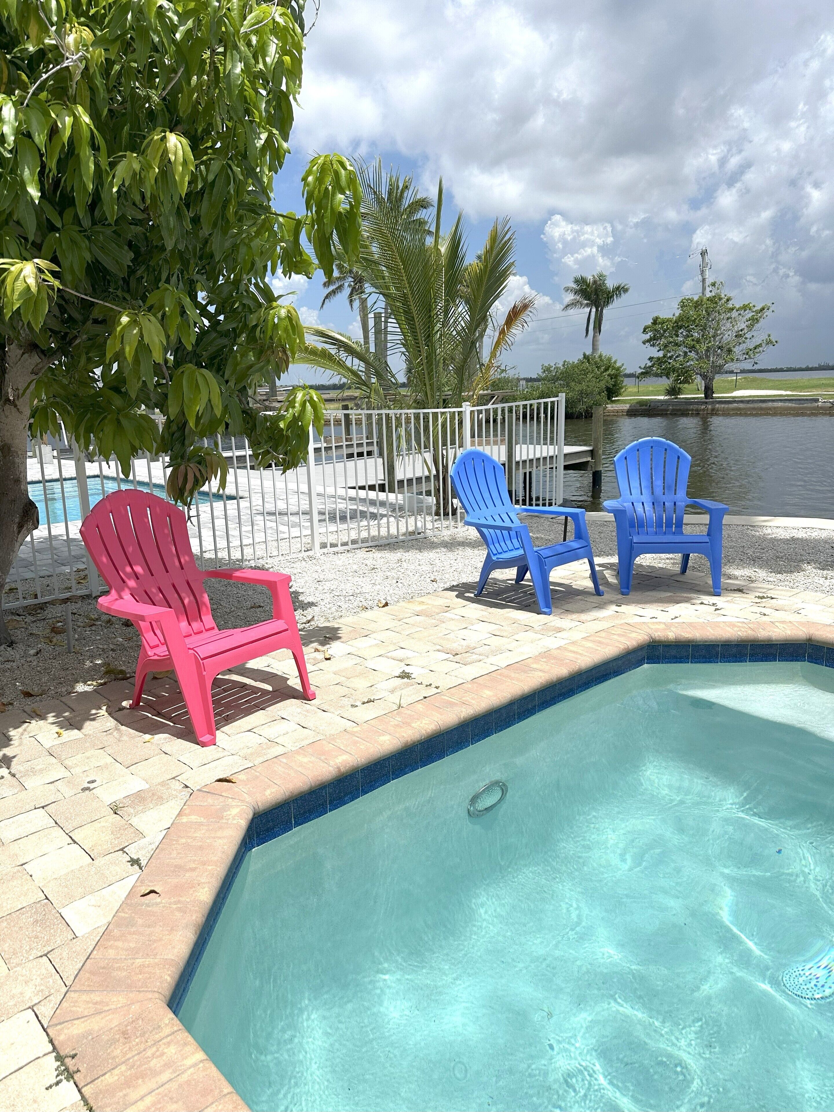 A pool area with a clear blue pool, surrounded by a white fence, and featuring three Adirondack chairs.