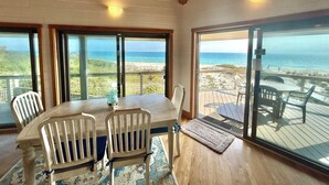 Un comedor con mesa y sillas, ventanas amplias con vista a la playa y al océano, y techo de madera.